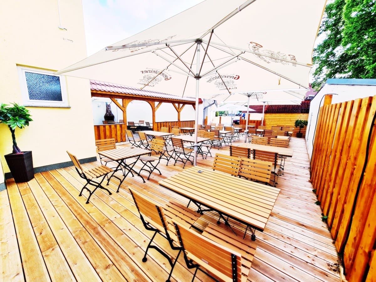 Biergarten auf einer hellen Holzterrasse: Biertischgarnituren und Klappstühle unter großen Sonnenschirmen, dahinter ein Holzpavillon