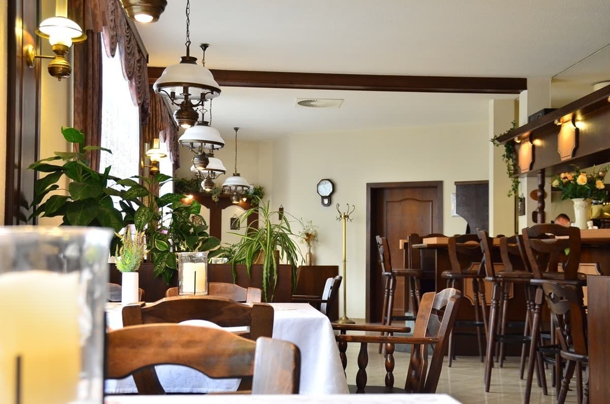 Gastraum des Restaurants: weiß eingedeckte Tische, dunkle Holzstühle, Hängeleuchten mit Milchglasschirmen und Grünpflanzen vor den Fenstern