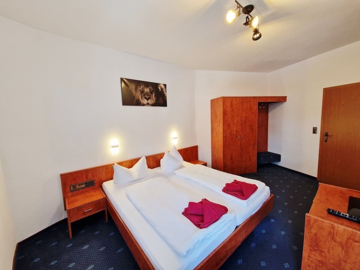 Doppelzimmer mit breitem Holzbett, weißer Bettwäsche und roten Handtüchern, Nachttisch, Kleiderschrank und dunkelblauem Teppichboden