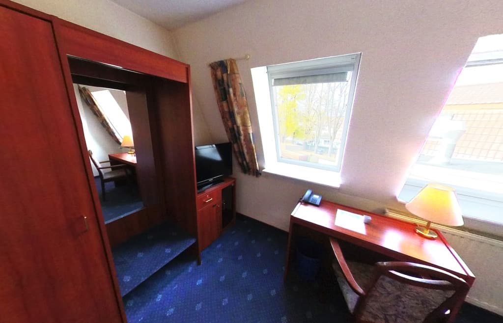 Zimmer unter der Dachschräge mit Schreibtisch, Tischleuchte und Telefon am Fenster, daneben Kleiderschrank und Fernseher