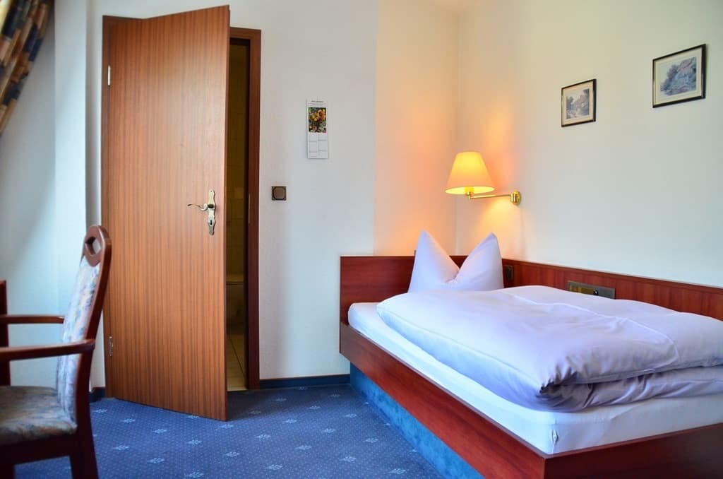 Einzelzimmer mit Bett, warmer Leseleuchte über dem Kopfteil, zwei kleinen Bildern an der Wand und offener Tür zum eigenen Bad